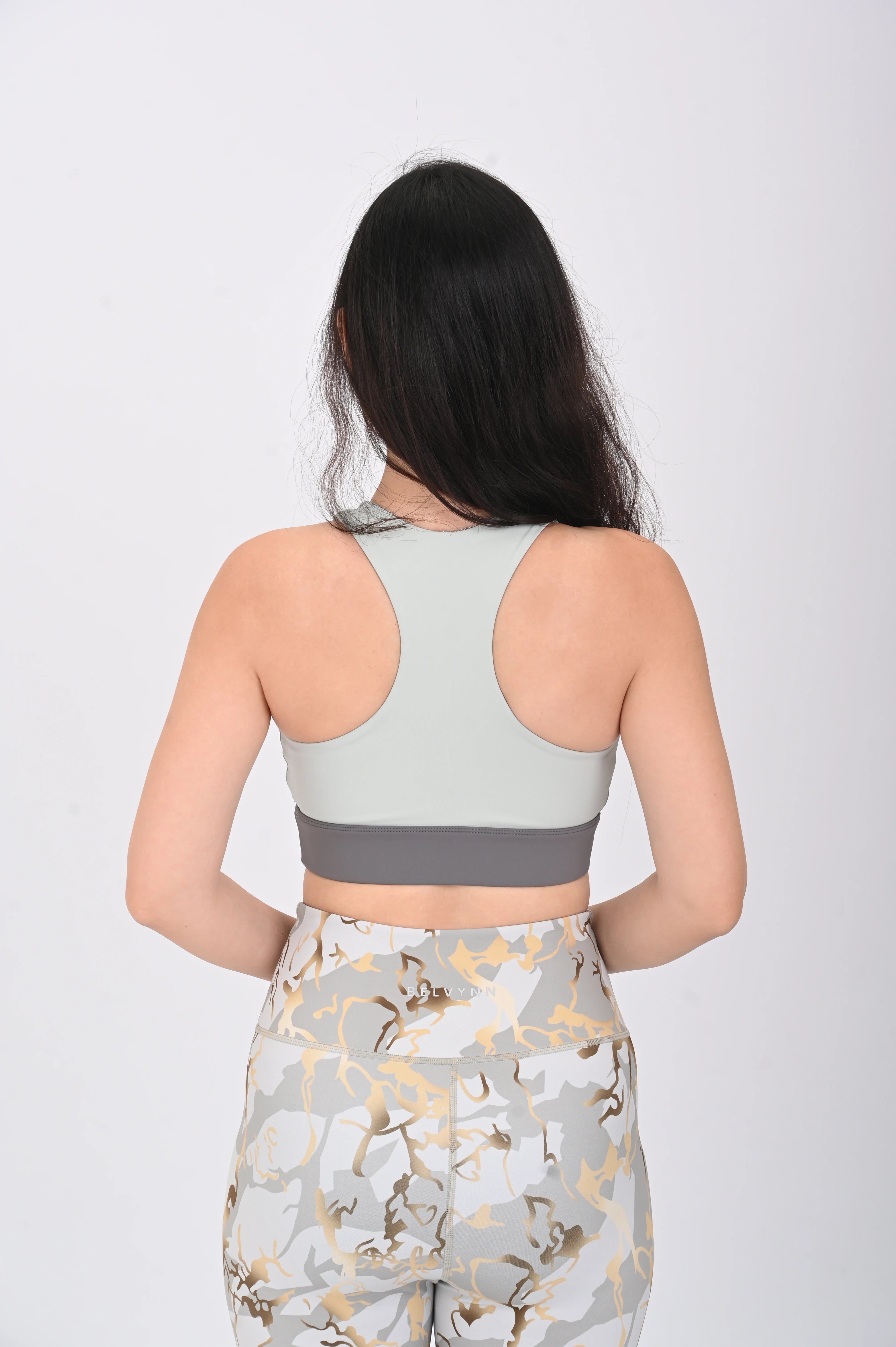 Back pose view of a Woman wearing a BELVYNN Signature ash green sports bra and floral leggings on a white background