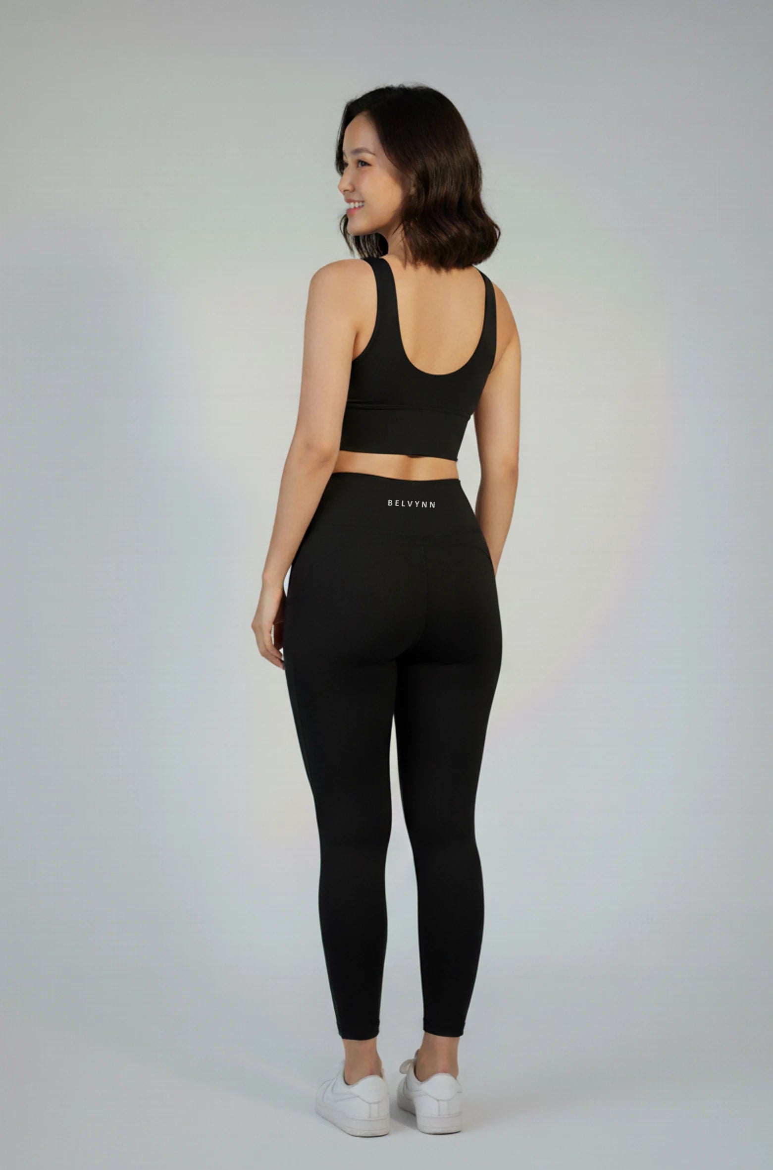 Back view of a woman wearing a BELVYNN premium black athletic bra and leggings with visible branding on a gray background