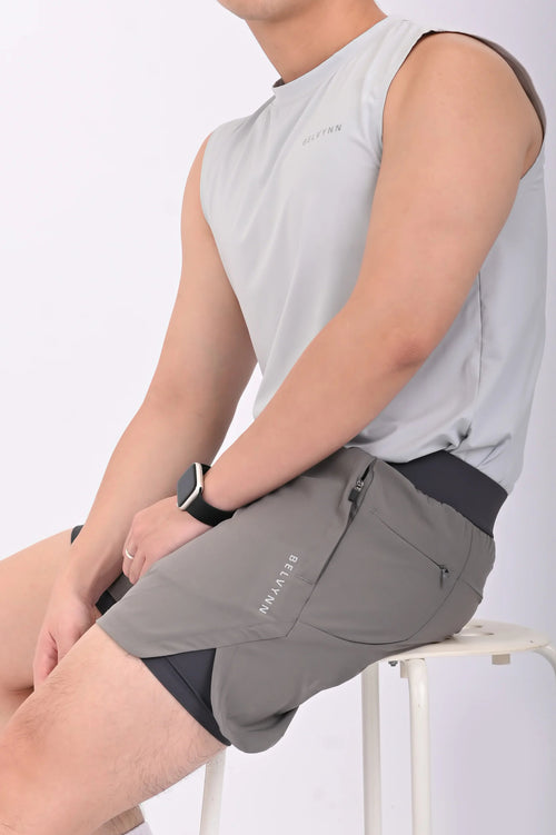 Person sitting on a white stool wearing BELVYNN premium gray shorts and gray tank top against a white background