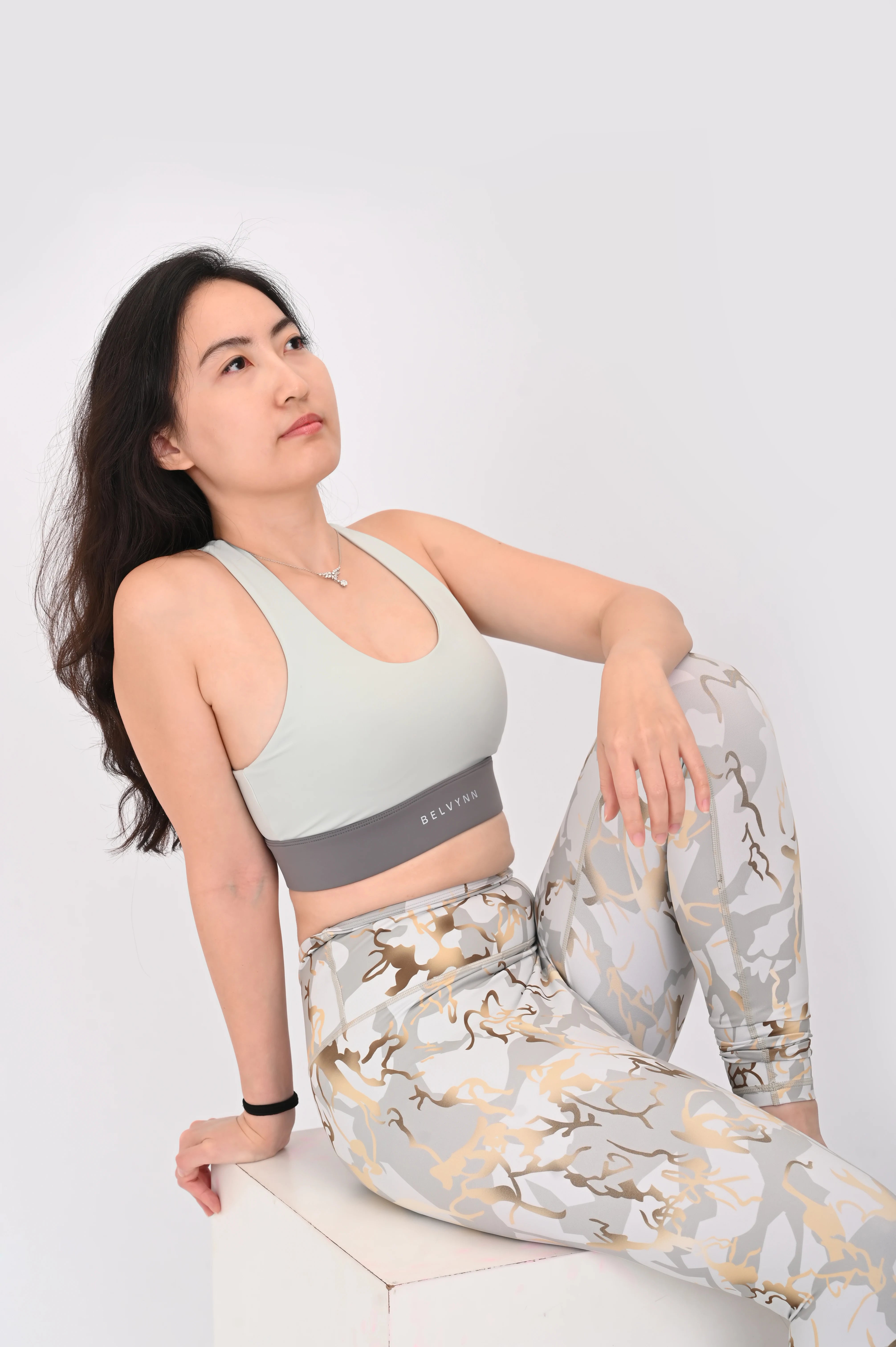 Relaxing pose of a woman wearing a BELVYNN signature light gray sports bra and patterned leggings on a white background