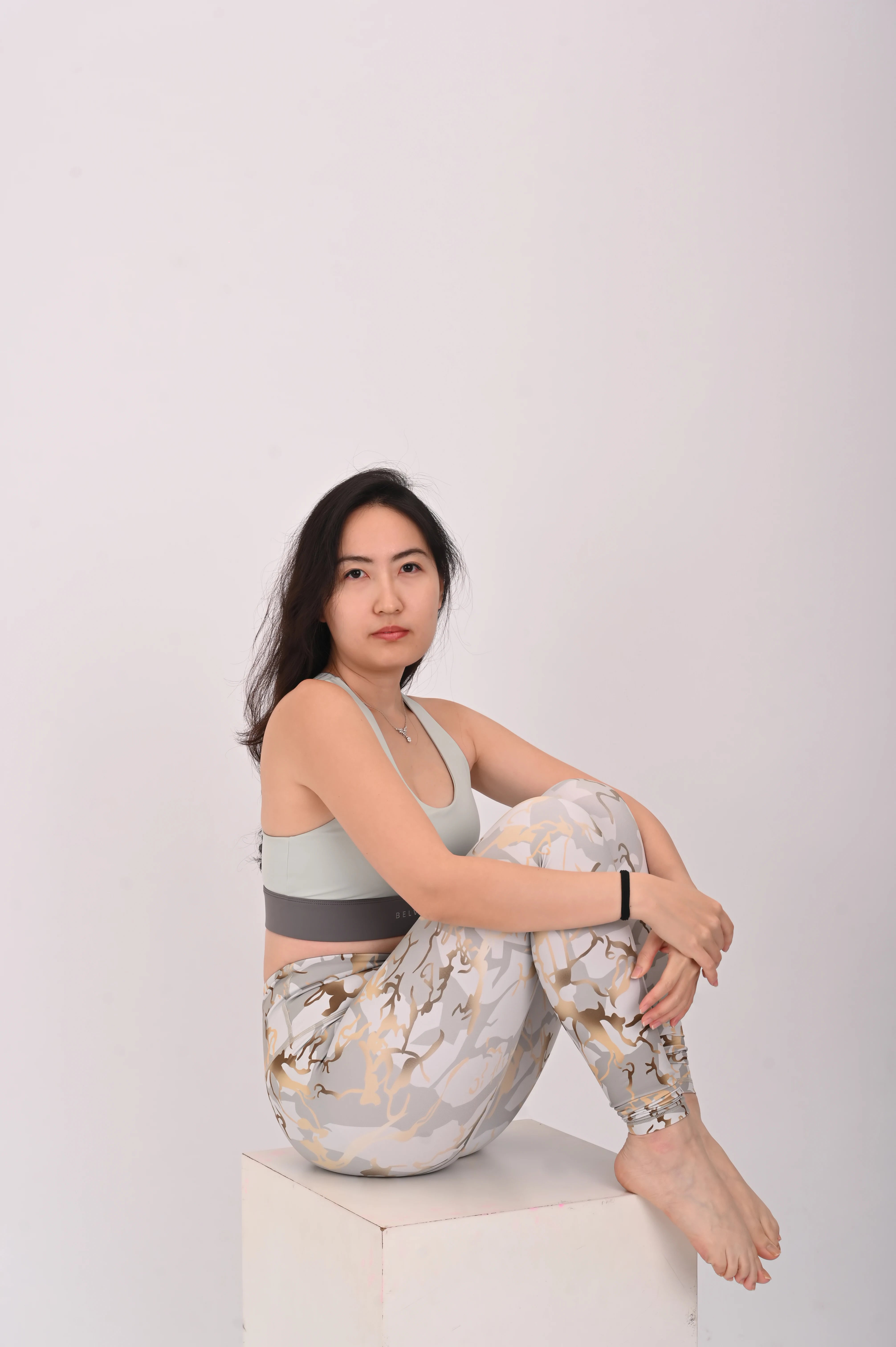 Woman sitting on a white box wearing a BELVYNN premium light gray sports bra and patterned leggings against a white background
