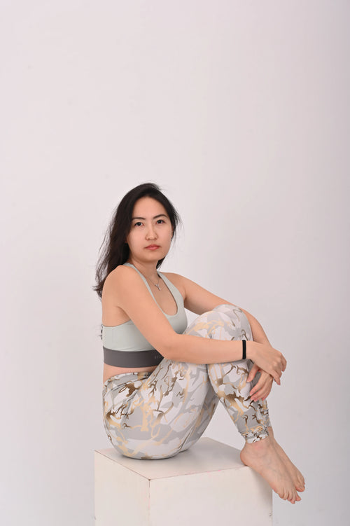 Woman sitting on a white box wearing a BELVYNN premium light gray sports bra and patterned leggings against a white background