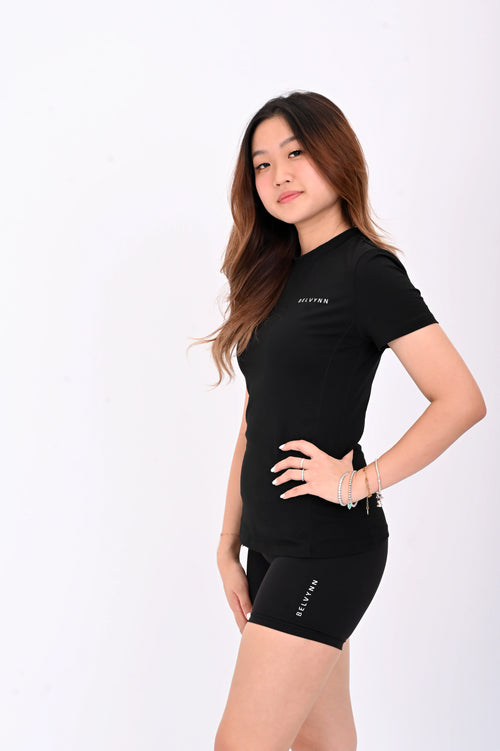 Woman standing and posing in a BELVYNN premium black athletic outfit with visible branding on a white background