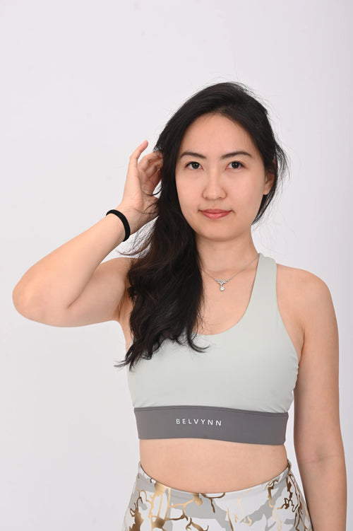 Woman wearing a BELVYNN Signature light gray sports bra with branding on a white background